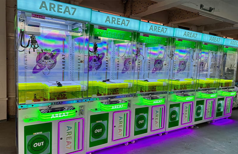 A row of neon green LED illuminated Area7 claw crane machines for plush toy vending in game centers.